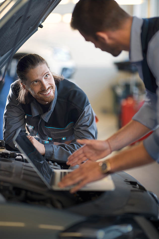 Zwei Kfz-Mechaniker in Uniform arbeiten in einer geschäftigen Werkstatt unter der Motorhaube eines Autos; der eine benutzt einen Laptop, während der andere zusieht und lächelt.