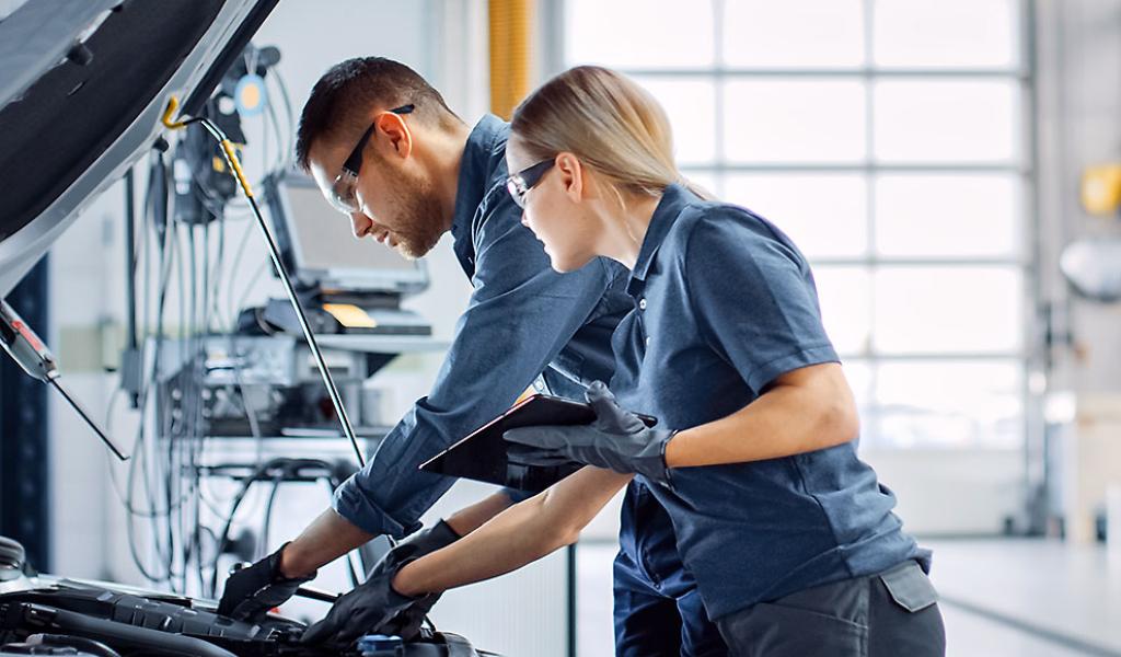 Zwei Kfz-Mechaniker, die Schutzbrillen und Handschuhe tragen, überprüfen in einer Werkstatt einen Automotor; einer kontrolliert die Teile, während der andere ein Klemmbrett in der Hand hält.